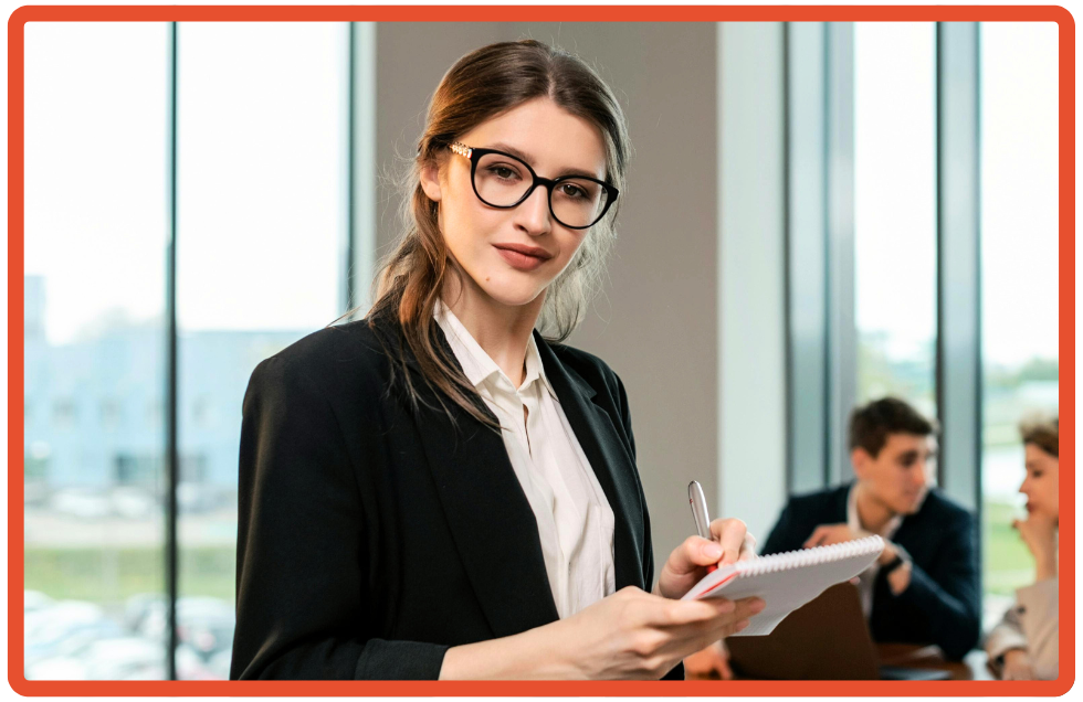 Professional woman in glasses and business attire holding a notepad and pen in a modern office conference room setting.