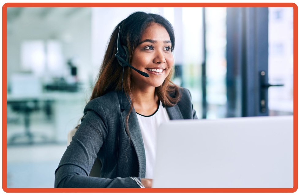 Smiling customer service representative wearing a headset and blazer, working at a laptop in a bright modern office.